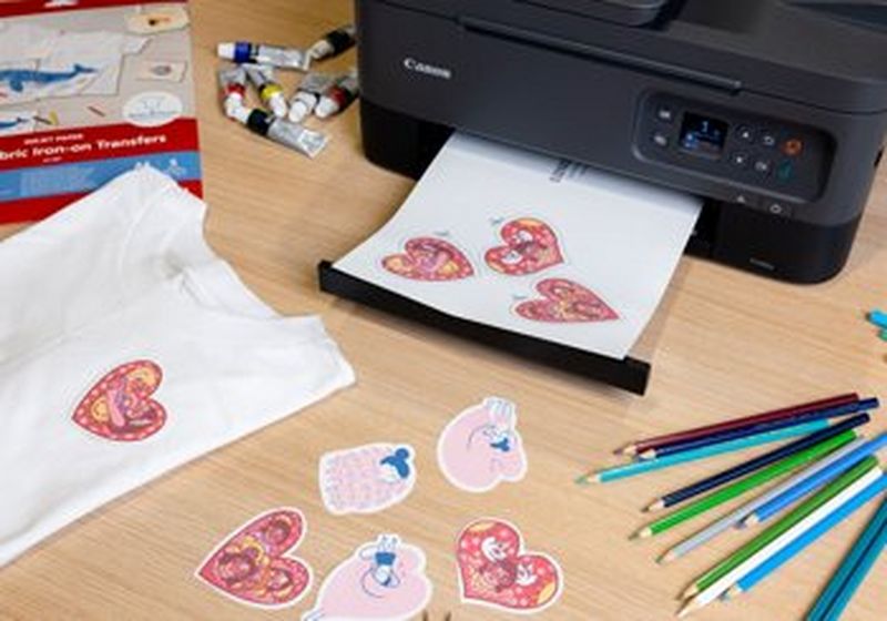 A Canon printer sits on a wooden desk. It has just printed iron-on transfers of three hearts and a further five more are sat on the desk in front of it. On the table there are also a folded white t-shirt with a heart illustration printed on the front left-hand side, some coloured pencils, seven small tubes of paint and a pack of Canon light fabric iron on transfer paper.