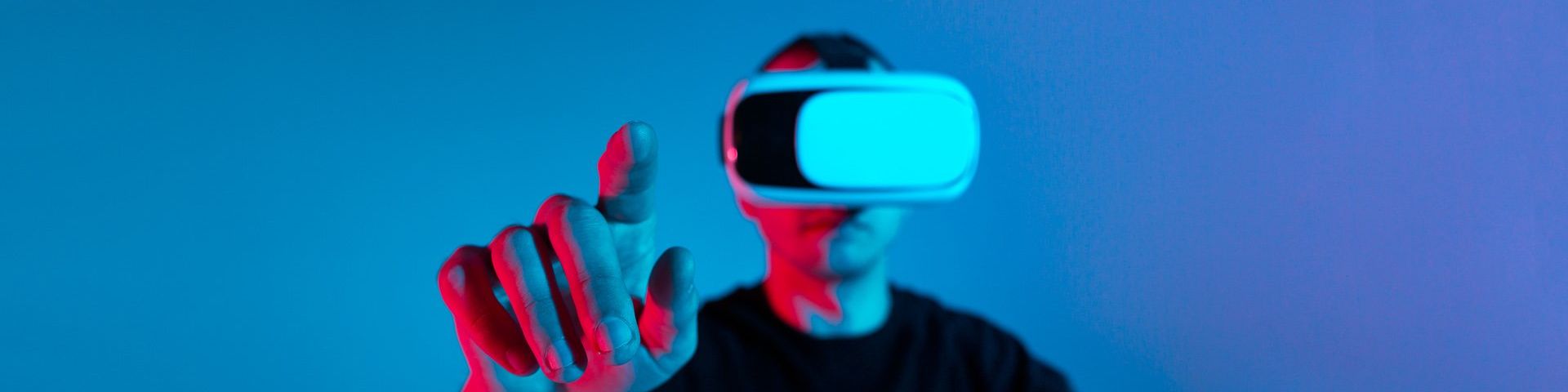 A man in a VR headset stands against a blue background with his finger pointed to the camera.