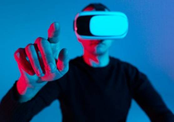 A man in a VR headset stands against a blue background with his finger pointed to the camera.
