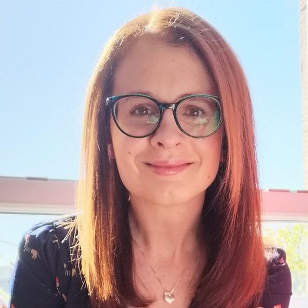 A smiling, red haired woman in glasses, sits in front of a window, through which you can see a bright blue sky. She wears a silver necklace with a pendant.