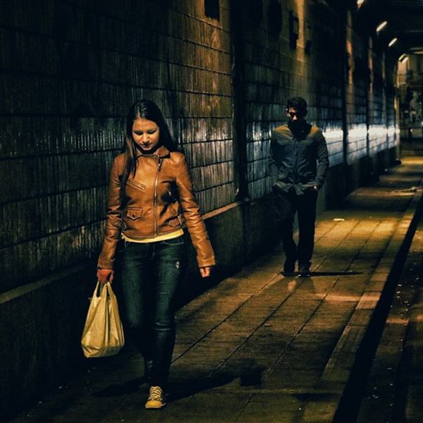 A young woman in a brown leather jacket, carrying a shopping bag, walks down a dark street at night. A short distance behind her, she is followed by a man, whose features are obscured by the dark. 