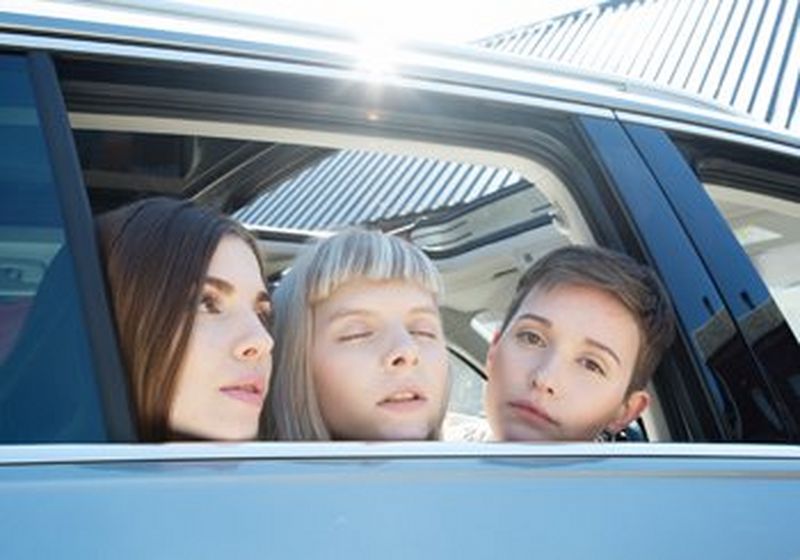 A close-up of three women leaning their heads out of a car window. The woman on the left has long brown hair, the woman on the right has short brown hair and, in the middle, the woman is blonde and has her eyes closed and head raised gently skywards.