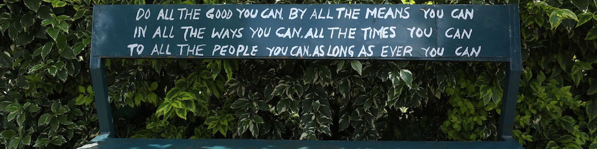 A green bench against a background of leaves. On the bench is hand painted in white letters: “Do all the good you can by all the means you can, in all the ways you can, all the times you can, to all the people you can, as long as ever you can.”