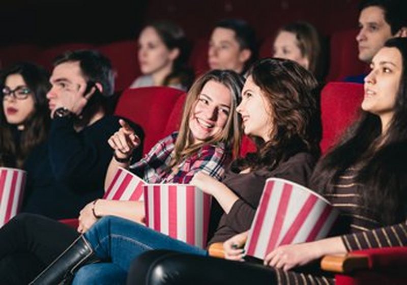 Two women sat in the cinema laugh together. One points towards the screen. They are holding red and white popcorn buckets and there are other cinemagoers around them. The chairs they sit on are red.