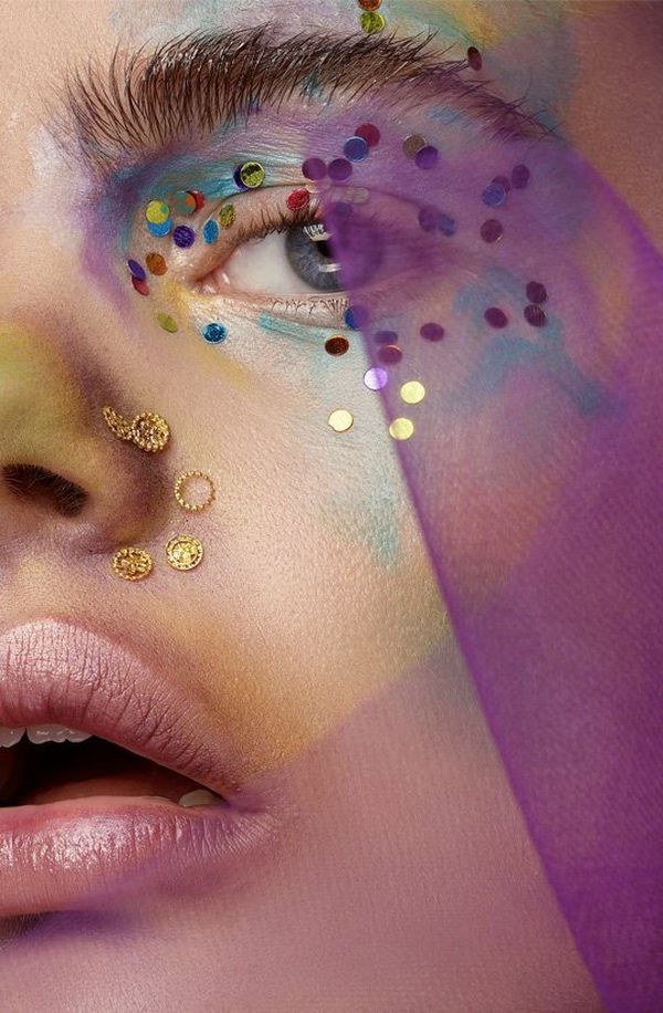 A close-up shot of a model’s face, which is partially covered in a sheer purple material. Her eyes are made up ‘watercolour style’ in pastel shades of purple, green and blue and she has gems alongside her nose.