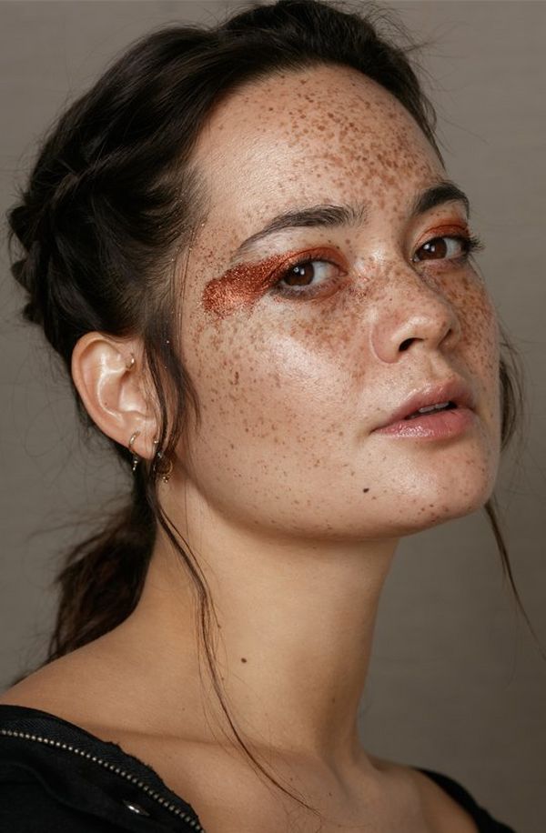 A model with very natural braided, dark hair wears a smear of bronze on her eyelids. The rest of her face is highly freckled and natural looking, but she has a dewy complexion with an attractive shine on her cheekbones, forehead and chin.