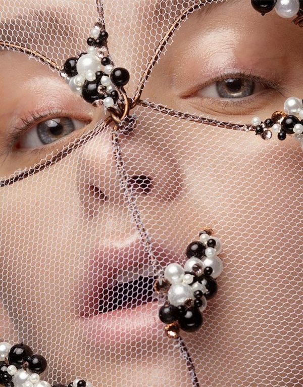 A model poses with her mouth open. Her face is partially obscured by a loose mesh, which has black and white pearl-like beads attached to it and holes for the model’s eyes. Her make-up is subtle, with a glow on her eyelids and upper lip.