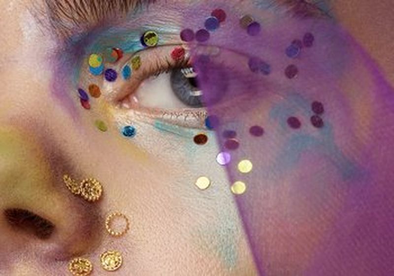 A close-up shot of a model’s face, which is partially covered in a sheer purple material. Her eyes are made up ‘watercolour style’ in pastel shades of purple, green and blue and she has gems alongside her nose.
