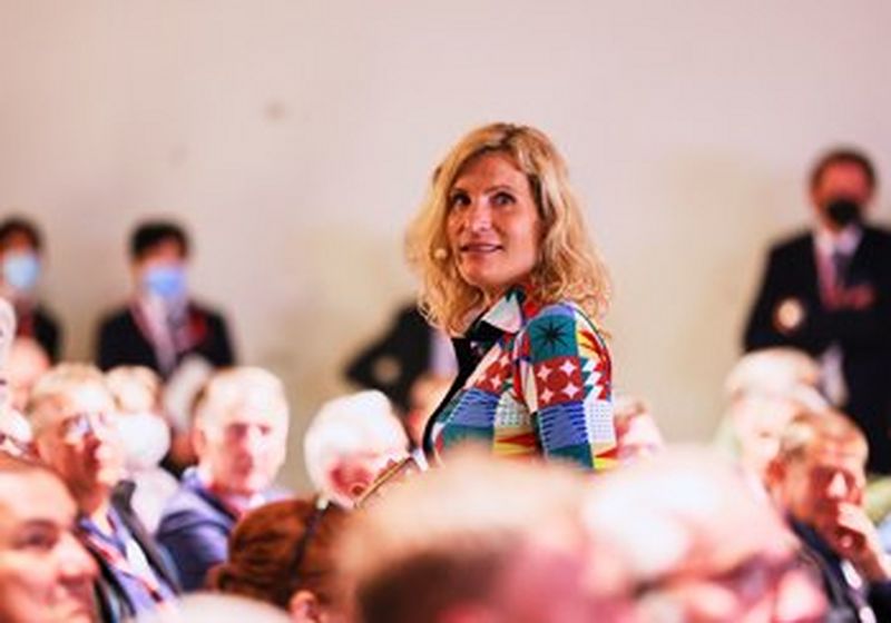 A blonde woman with shoulder length hair and a headset mic, surrounded by seated and standing men, whose faces are largely blurred. Only her upper body can be seen, but she is wearing a multicoloured dress of blocks of different patterns – stars, zigzags and triangles.