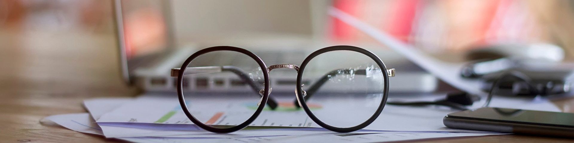 A pair of spectacles sat on top of paperwork, in front of a laptop and next to a phone