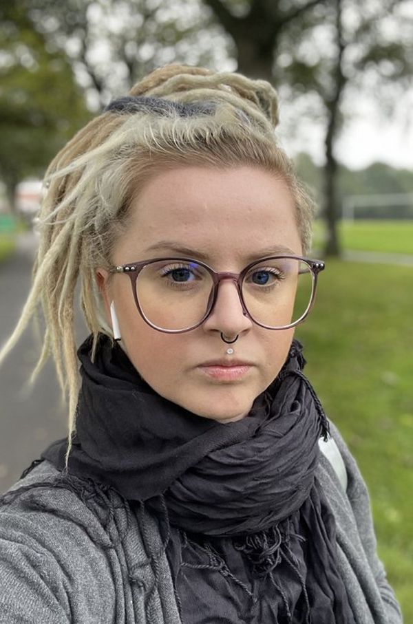 A blonde haired woman with glasses wearing a black scarf and grey top. She has a white headphone visible in her right ear and has a piercing through her septum and philtrum.