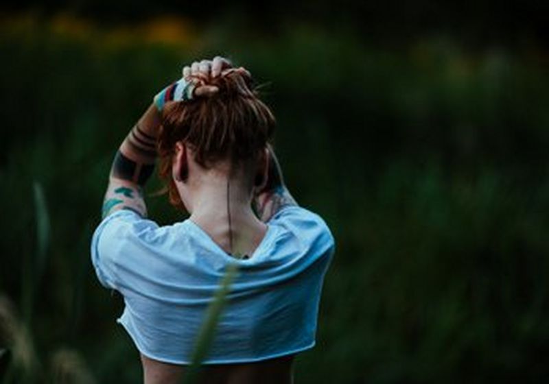 A person in a light-coloured t-shirt, photographed from the back, against a natural green background. They have their tattooed arms held in the air, above their head, as though holding their red hair up. © Marta Sobilo