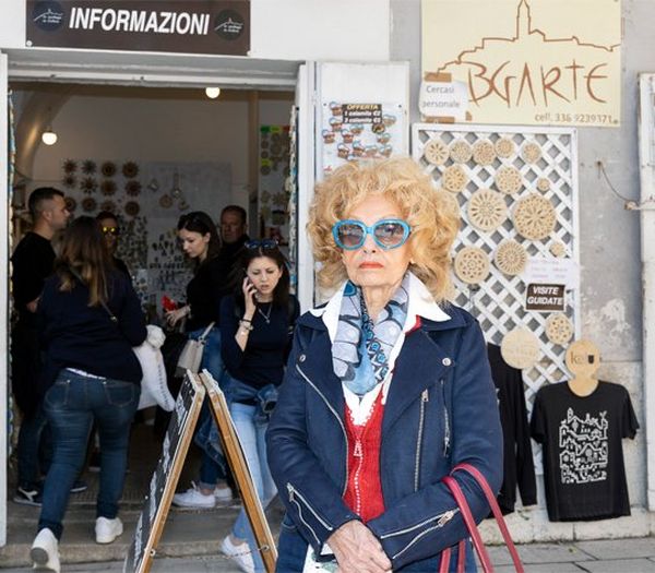 An older woman wearing with short blonde curly hair and large blue sunglasses and a blue biker-style jacket stands on the street in front of a group of young people in a doorway. 