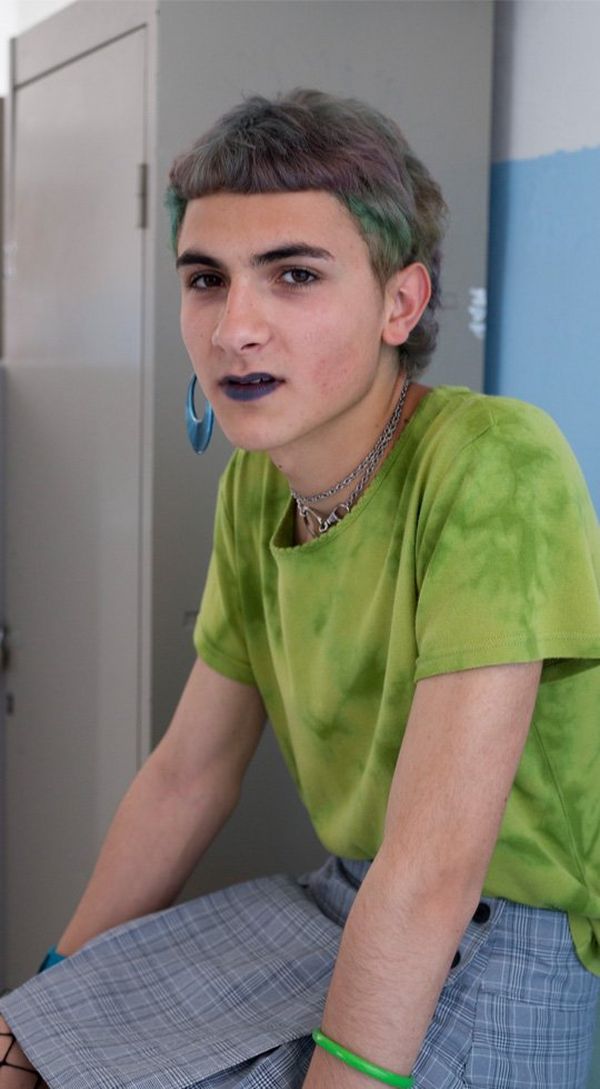 A young person with dyed green, grey and blue hair, wearing a green t-shirt and checked kilt.