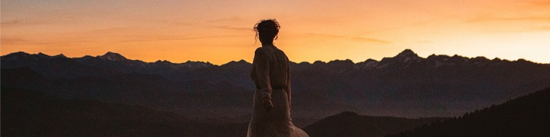 A woman stands with her back to the camera, watching an orange sunset over dark mountains. She wears a pale dress, and her hair is worn up, with tendrils outlined in shadow.