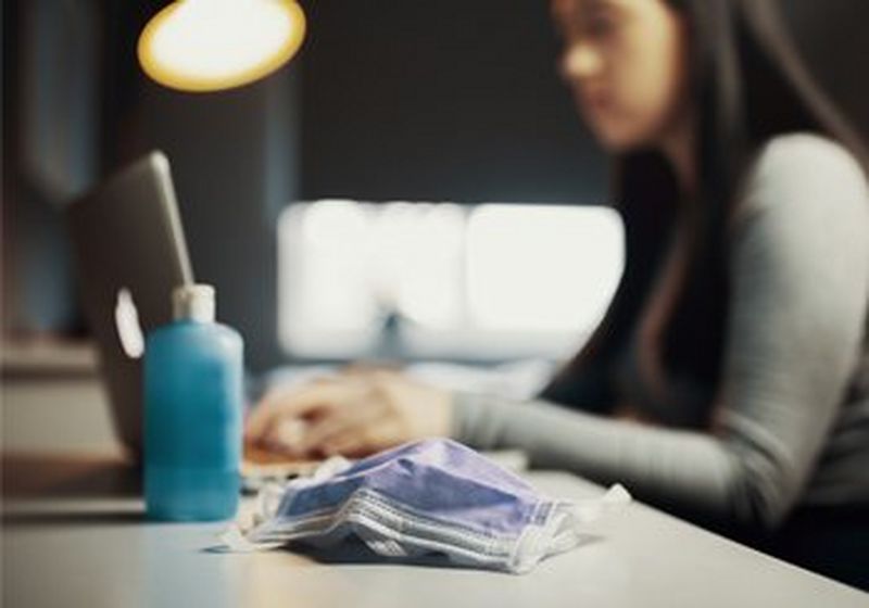 A medical face covering sat on a white office desk beside what appears to be a blue bottle of hand sanitising gel. Behind these is the upper body of a person sat at the desk, wearing a long-sleeved grey top and typing on a laptop.
