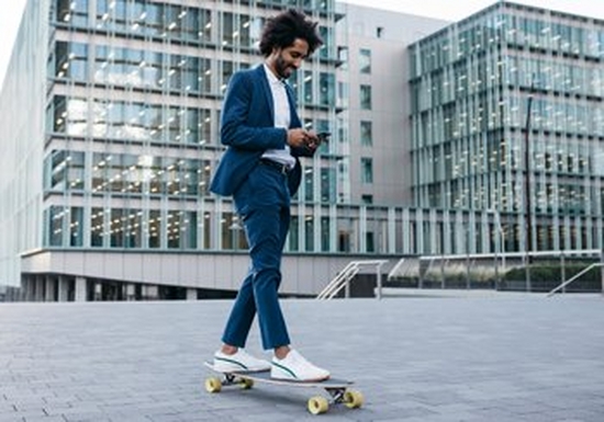 A man wearing a blue suits and white shirt is on a skateboard, checking his phone. Behind him are glass fronted offices
