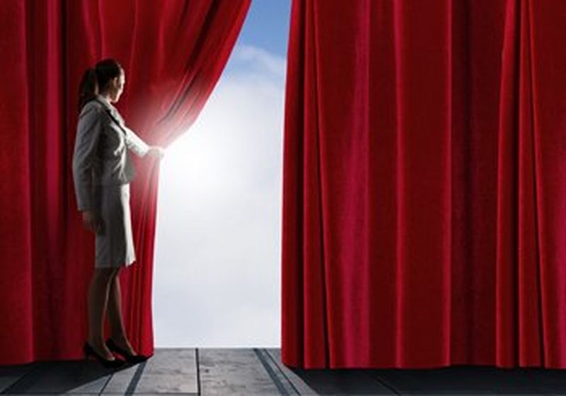 A woman wearing a business suit pulls back a red curtain to reveal a bright blue sky and clouds.