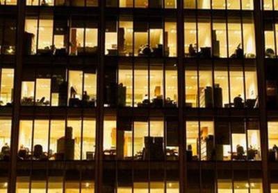 A view into an office building at night, with the windows lit up in yellow and office equipment in silhouette.