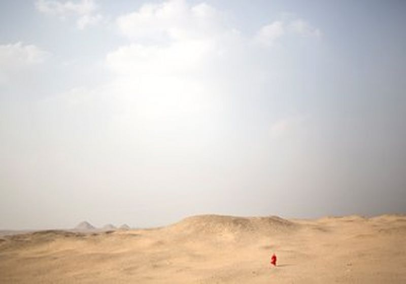 A women in a red dress is walking in the desert. She is tiny and the desert is massive as the sand stretches far.