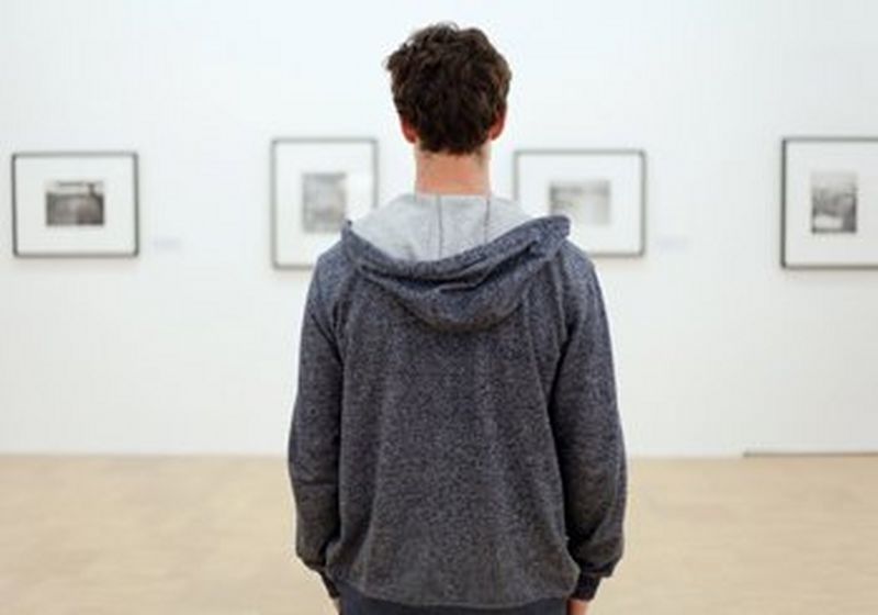 A person with short dark hair and wearing a grey hoodie stands with their back to the camera. They are facing four out of focus pictures in black frames with white mounts, hanging on a white gallery wall.