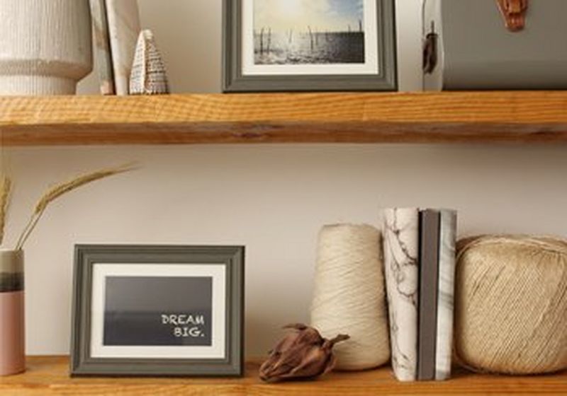 Office shelves with a picture frame containing the quote ‘dream big’, around it are a dried flower, a ball of thread, some notebooks, pots and another frame with a seascape photo. 