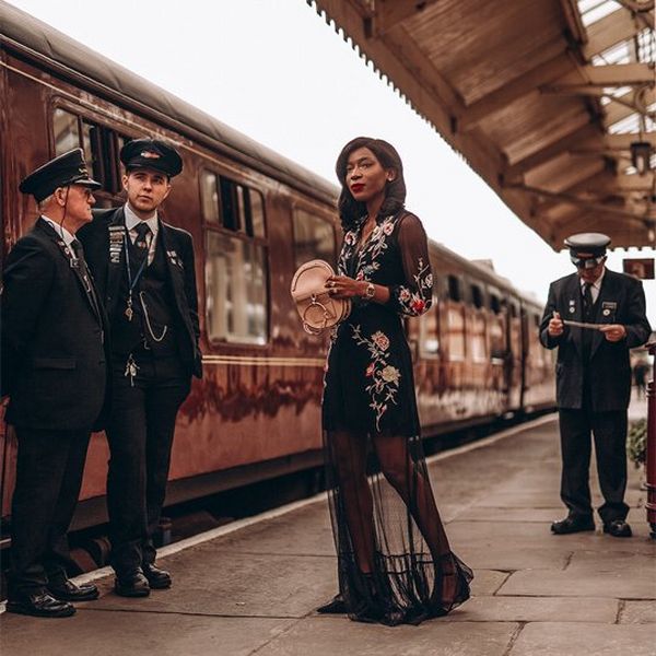 A woman stands on a train platform, wearing a black dress, with pastel-coloured floral trims and holding a small pink handbag, as though reaching inside. An old-fashioned red train sits on the tracks and three men in vintage conductors’ uniforms stand nearby.