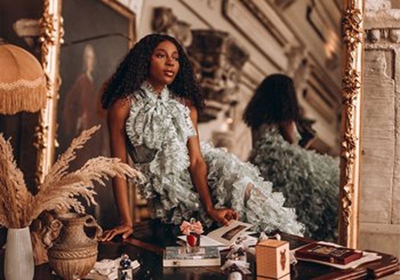 A woman sits on a desk in front of a huge ornate, rose gold mirror, in which the room is reflected. She is wearing a mint green halter neck dress with ruffles. In front of her, on the desk, is an assortment of objects – vases, with dried flowers, books, perfume bottles and small glass objects.