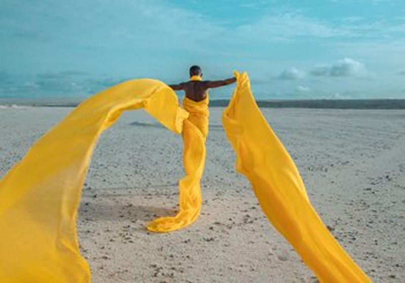 A woman walks on a sandy, flat surface towards the ocean. Her arms are stretched wide, and she wears bright yellow chiffon that trails behind her, blowing in the wind and creating a strong contrast to the blue sky.