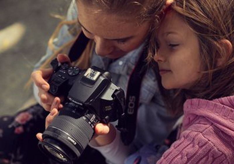 A photo taken from above of two children – one in a blue denim jacket, the other in a pink knitted top. One child holds a large Canon camera in their hands, with the strap around their neck. They both peer, smiling, at the screen on the back of the camera. 