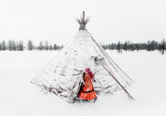 	The image is dominated by pure white snow, save a snow-covered chum, or traditional tent used by the migrational tribes of the region. A woman emerges from her tent and is wearing a long orange/red patterned dress and boots. The dress has a black trim midway up the skirt and she also wears a pink headscarf, tied at the back of her head, which swings behind her.