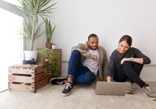 Two casually dressed young people sit on the carpeted floor of a white walled room, next to some crates with plants on them. They are both looking at a laptop.