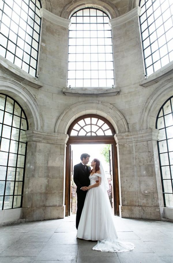 Newlyweds stand in front of the door of the grey stone atrium of a church, surrounded by huge arched leaded windows.