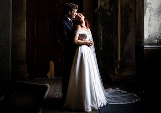 An immaculate bride and groom stand in a shabby room with the light pouring onto the bride's white dress. She stands with her back to her husband and his arms around her waist, and looks back and up into his eyes.  