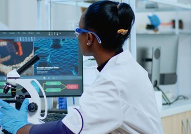 A female clinician in a short sleeved white lab coat, wearing latex gloves sits at a microscope with a screen beside it. The purple sleeves of her t-shirt show and she wears lab goggles with blue arms. Her hair is in a bun. On the screen are medical images of what she is viewing.