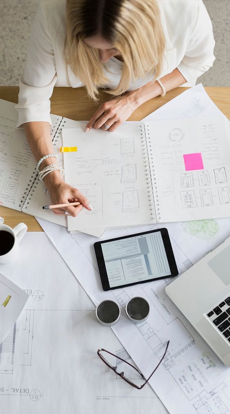 Overhead shot of a blond woman in a white shirt sitting at a desk covered with a laptop, tablet device, and papers, working in a ring-bound book.