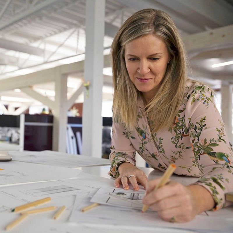 Woman with dark blond hair leans over a large white desk with laptop and papers, writing on a page with coloured pencils. 