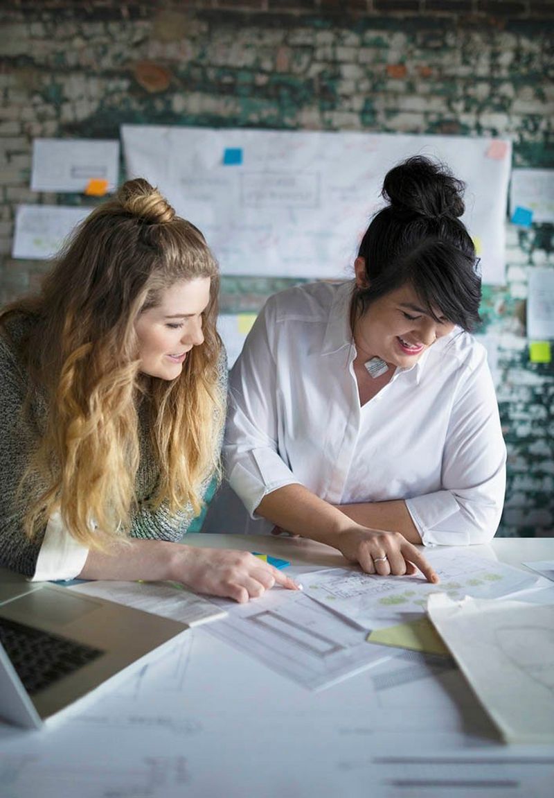 Young woman with long wavy blonde hair and middle-aged woman with dark hair in a bun look at papers spread out on a white table in front of an exposed brick wall.