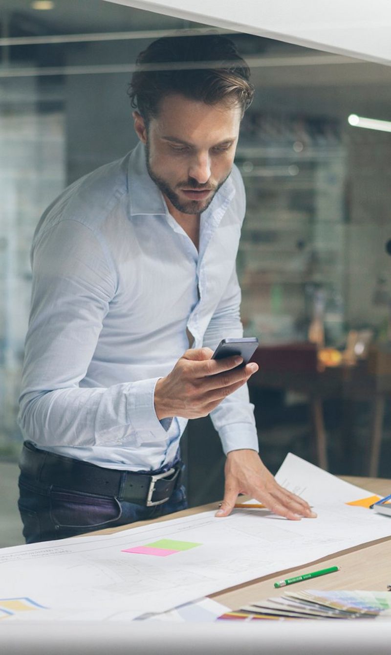 Man with beard wearing light blue shirt leans over a desk spread with papers and a model of a building, looking at a smartphone. 
