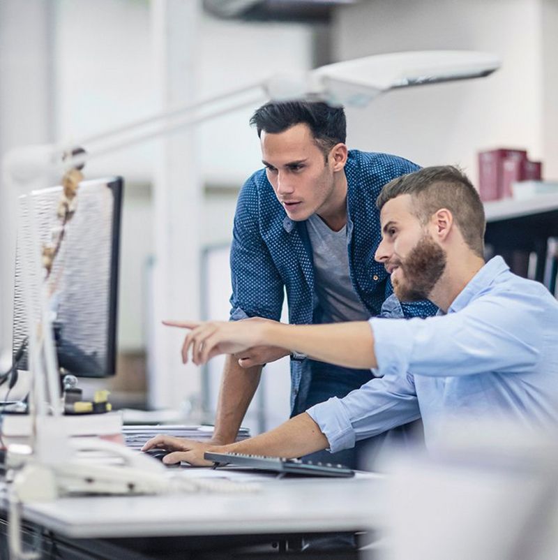 Two men wearing blue shirts, one with a beard, crowd round a desk, pointing at a large computer screen.