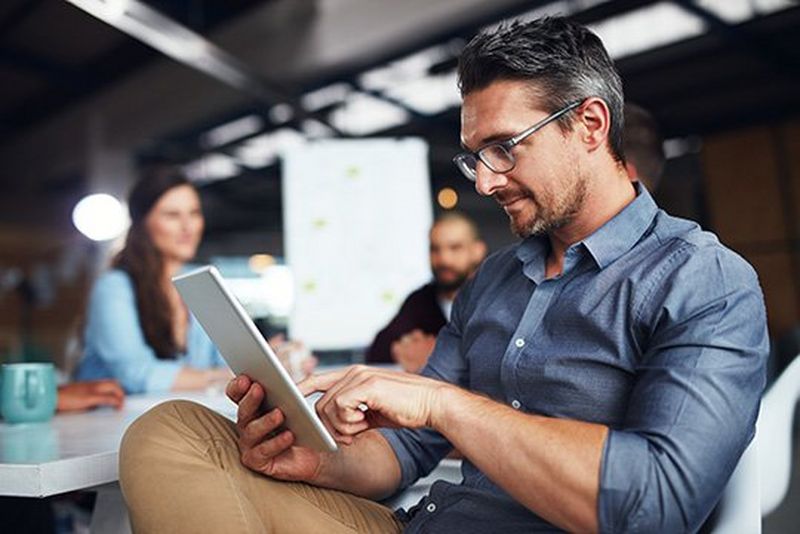 Man with glasses and stubble wearing a blue shirt and beige chinos sits at a desk and uses a finger to scroll down a tablet device.