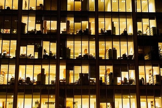 A view into an office building at night, with the windows lit up in yellow and office equipment in silhouette.