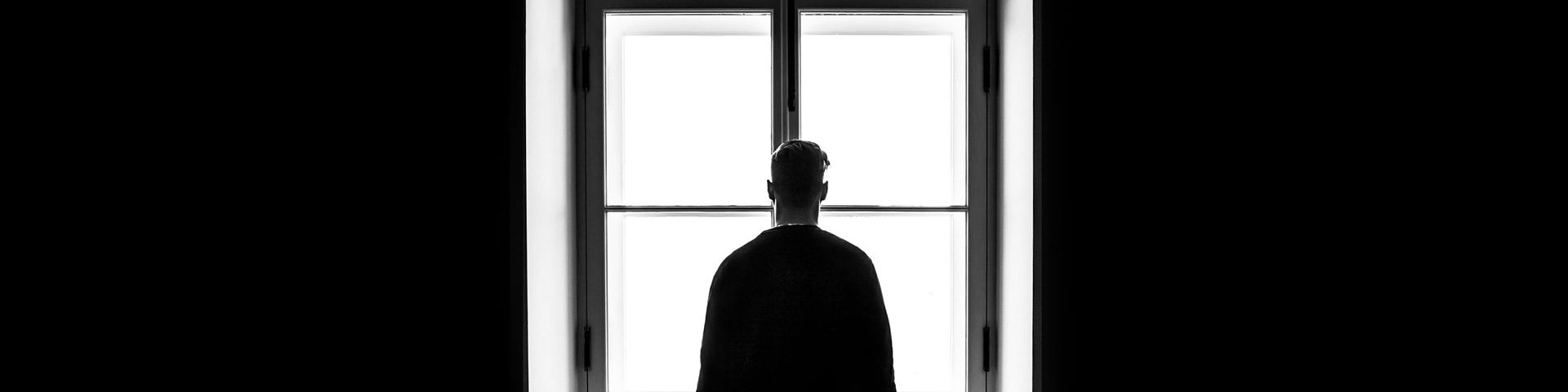 A black and white photograph of a back of a man, silhouetted, stood in front of at the bright white light of window. On either side, the walls are black. 