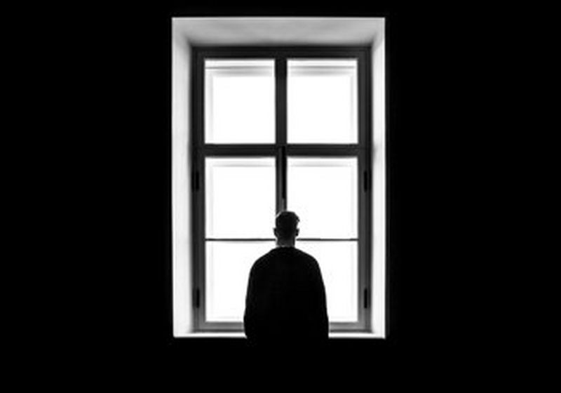 A black and white photograph of a back of a man, silhouetted, stood in front of at the bright white light of window. On either side, the walls are black. 