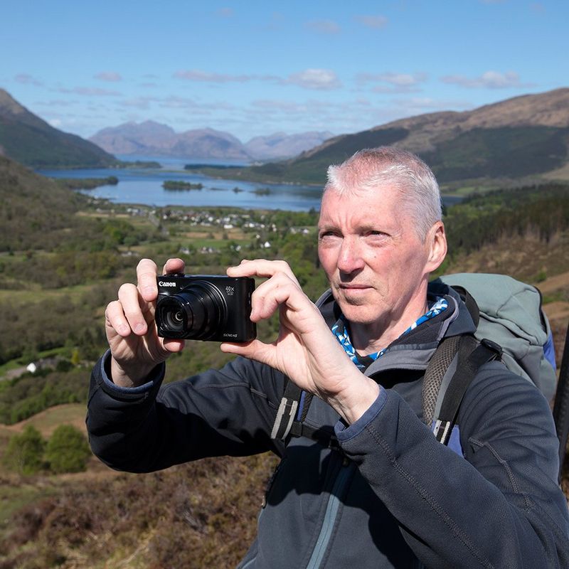 Alan Rowan stands on the side of a hill, with yellow gorse bushes, grassy fields and a river in the valley behind him. He holds a compact camera to take a photo of the natural view.