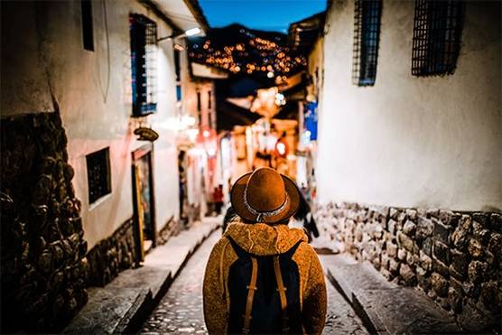 A tourist, wearing a coat, a backpack and a hat, pictured from behind facing an old cobbled street at evening with lights twinkling on a dark hill in the background. 