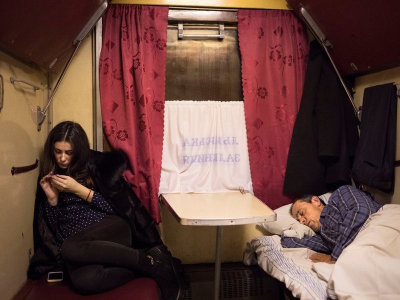 Two passengers in a compartment on a train. One is lying down sleeping, the other sits curled up on her seat inspecting a fingernail.