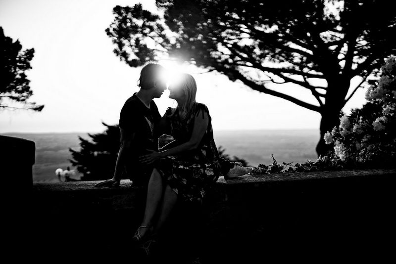A couple sitting together on a wall facing each other, with the light of the sun shining between their heads.