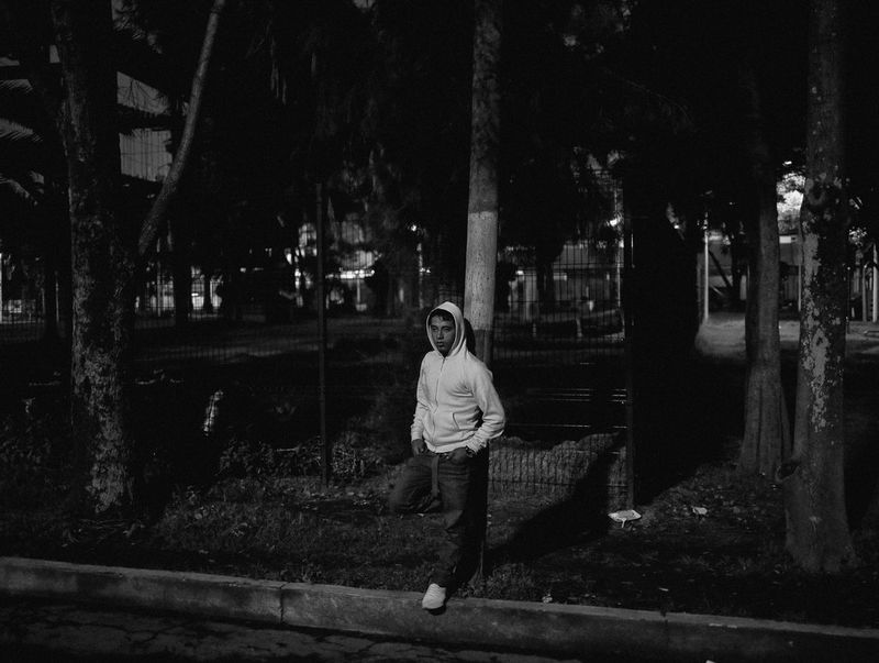A youth wearing a hoodie leans against a lamppost in front of a tree-filled urban park in the evening.