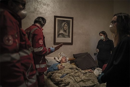A man lies in bed surrounded by Italian Red Cross volunteers. A painting of the Virgin Mary hangs on the wall. Photo by Fabio Bucciarelli.